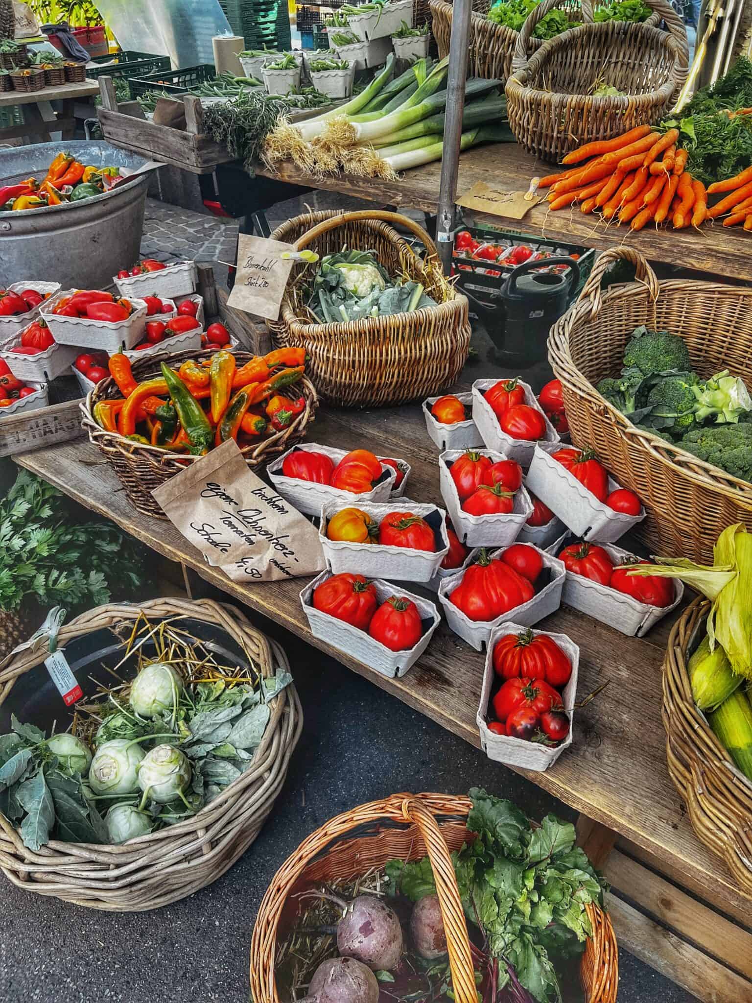 Frisches Bio-Gemüse auf Markt, bestehend aus Tomaten, Paprika, Brokkoli, Sellerie, Zwiebeln und Wurzeln, umgeben von handgefertigten Körben und Holztischen.