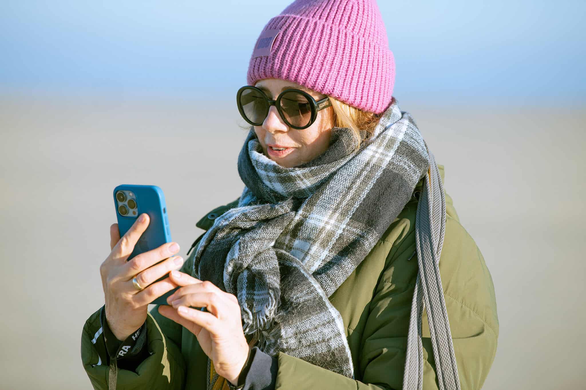Frau in Mütze, Sonnenbrille und Schal nutzt ihr Handy am Strand, urbaner Lifestyle, Selbstreflexion, authentisch, outdoor, moderne Frau, Mode, Natur, Technik, Selfcare, Lifestyle, Selbstentwicklung.